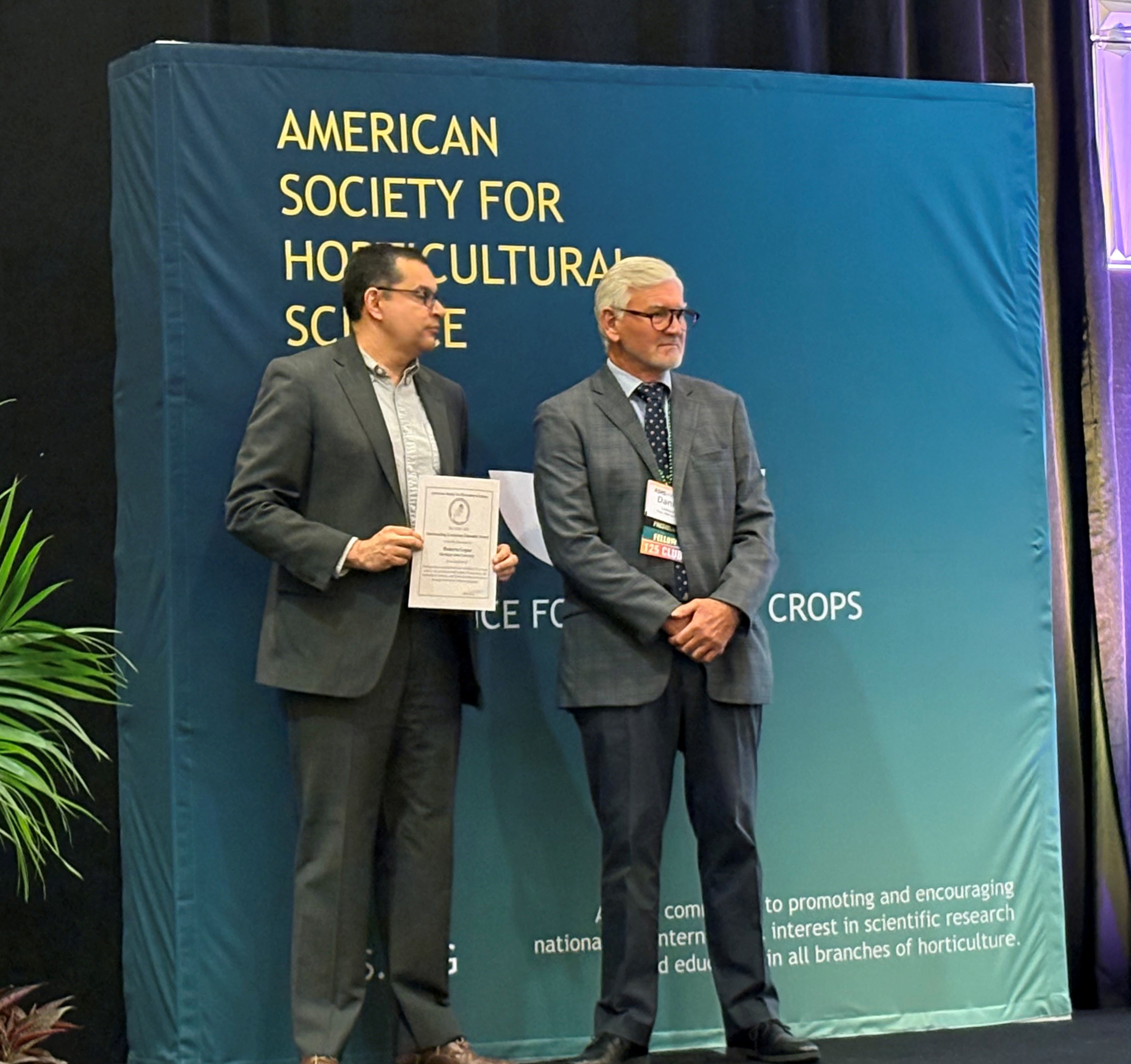 Roberto Lopez, wearing a suit, stands on stage holding an award certificate next to another man in a suit at the American Society for Horticultural Science annual conference.